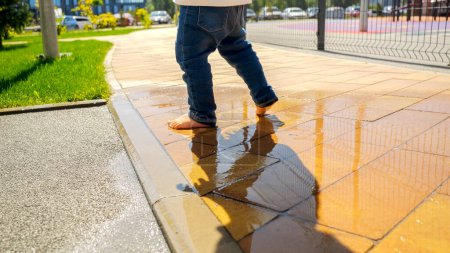Photo for Closeup of barefoot toddler boy walking over wet water puddle on pathway in park or playground. - Royalty Free Image