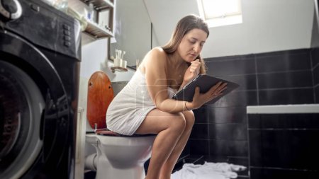 Foto de Una mujer que trabaja en una tableta mientras está sentada en un inodoro. La imagen enfatiza el concepto de trabajo remoto, educación y ser un individuo dedicado - Imagen libre de derechos