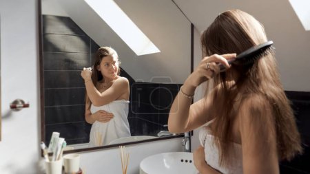 Foto de Mujer de pie en un baño, cubierta con una toalla, y peinándose el pelo largo mientras se mira en el espejo. Concepto de mantener una buena higiene y rituales de autocuidado - Imagen libre de derechos