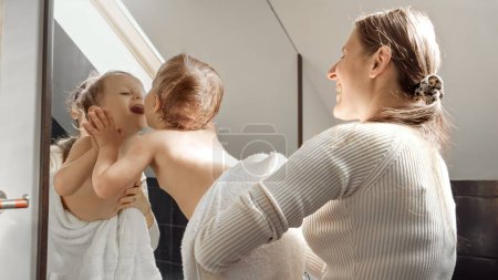 Téléchargez les photos : Bébé garçon mignon couvert de serviette de bain avec la mère regardant dans son reflet dans le miroir. Concept de parentalité, bonheur et hygiène de l'enfant - en image libre de droit