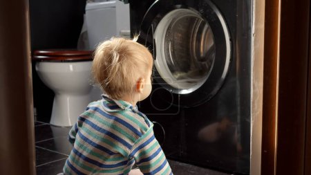 Téléchargez les photos : Bébé garçon souriant mignon regardant les vêtements de filature dans la machine à laver. Travaux ménagers et tâches ménagères, éducation et développement des enfants - en image libre de droit
