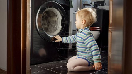 Photo for Portrait of baby boy watching washing machine spinning its drum. Doing housework and chores, children education and development - Royalty Free Image