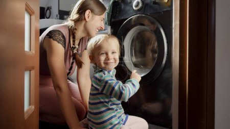 Foto de Madre feliz con el niño haciendo la colada y mirando la lavadora. Hacer tareas domésticas y tareas, educación y desarrollo de los niños - Imagen libre de derechos