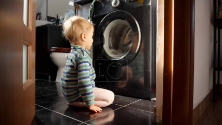Photo for Cute baby boy looking at washing machine doing laundry. Doing housework and chores, children education and development - Royalty Free Image