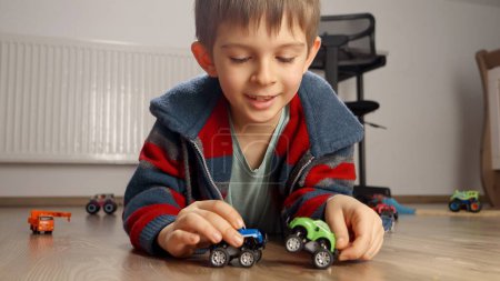 Foto de Retrato de niño lindo tirado en el suelo y jugando con dos coches de juguete. Niños jugando solos, desarrollo y educación, juegos en casa - Imagen libre de derechos