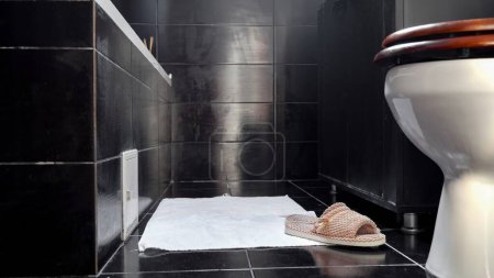 Foto de Zapatillas y toalla blanca tumbada en el suelo en el baño con azulejos negros. - Imagen libre de derechos