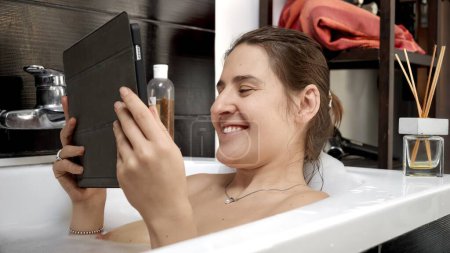 Photo for Woman relaxing in bath while having a video chat with friends or family on her tablet computer. Modern ways we balance personal care and social interaction - Royalty Free Image