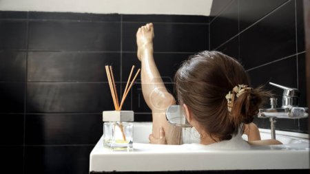 Téléchargez les photos : Une femme prenant un bain relaxant et se lavant les jambes avec de la mousse de savon. Importance de l'autosoin et de la relaxation. - en image libre de droit