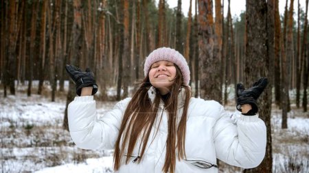 Téléchargez les photos : Portrait d'une adolescente souriante debout les yeux fermés dans la forêt d'hiver et attrapant des flocons de neige. - en image libre de droit
