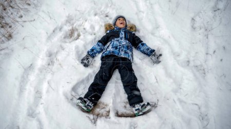 Téléchargez les photos : Petit garçon s'amuser dans la neige couché et faire ange de la neige. Personnes jouant à l'extérieur, vacances d'hiver et vacances, loisirs actifs - en image libre de droit