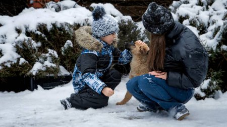 Foto de Pequeño niño y adolescente divirtiéndose con lindo perro cachorro en casa patio trasero cubierto de nieve. - Imagen libre de derechos