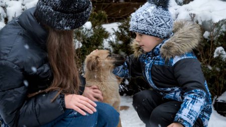 Foto de Lindo chico con chica adolescente acariciando y jugando con el perro durante las nevadas al aire libre. Niños con animales, juegos con mascotas - Imagen libre de derechos