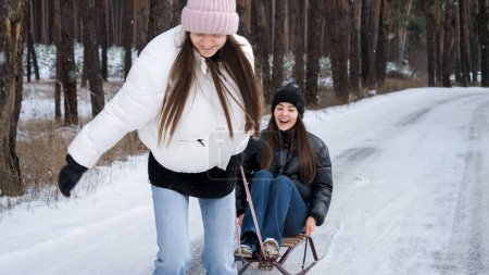 Photo for Two laughing teenage girls laughing and having fun while riding on sleighs at forest. People playing outdoors, winter holidays and vacation, active leisure - Royalty Free Image