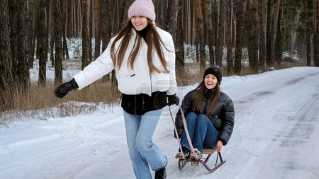 Téléchargez les photos : Deux adolescentes gaies chevauchant sur des traîneaux et s'amusant dans la forêt d'hiver. - en image libre de droit
