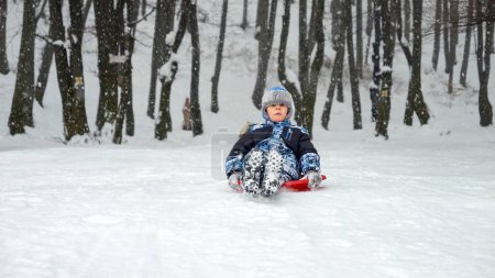 Téléchargez les photos : Garçon glissant doucement sur une colline enneigée sur son traîneau en plastique, souriant d'oreille en oreille. L'image parfaite des vacances d'hiver, du plaisir dans la neige et de la joie de l'enfance - en image libre de droit