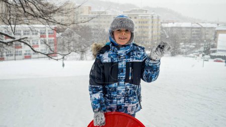 Téléchargez les photos : Joyeux garçon souriant tenant ses traîneaux en plastique sur une colline enneigée lors de chutes de neige. Le concept des enfants qui s'amusent en hiver, pendant les vacances de Noël et qui jouent dehors dans la neige - en image libre de droit