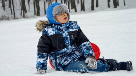 Téléchargez les photos : Petit garçon allongé sur la neige et frottant sa jambe blessée après être tombé. Blessures et traumatismes pendant les vacances d'hiver actives et les sports extrêmes. - en image libre de droit