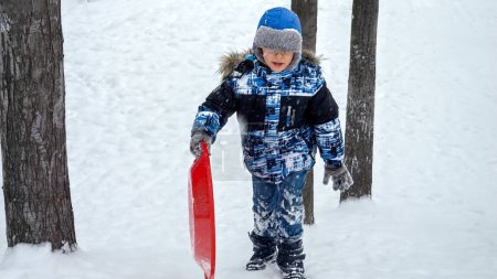 Téléchargez les photos : Joyeux garçon souriant escaladant une colline enneigée avec ses traîneaux en plastique lors de chutes de neige. Le concept des enfants qui s'amusent en hiver, pendant les vacances de Noël et qui jouent dehors dans la neige - en image libre de droit