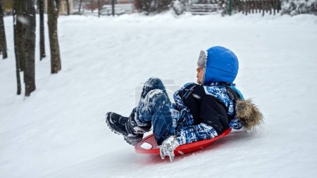 Photo for Cheerful boy riding down the snowy hill on plastic sleds during snowfall. Concept of winter holiday, children having fun and playing outdoors in the snow, Christmas vacation - Royalty Free Image