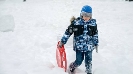 Téléchargez les photos : Joyeux garçon souriant marchant sur la colline enneigée avec ses traîneaux en plastique lors de chutes de neige. Le concept des enfants qui s'amusent en hiver, pendant les vacances de Noël et qui jouent dehors dans la neige - en image libre de droit