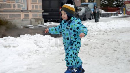 Téléchargez les photos : Joyeux petit garçon souriant qui court dans la rue enneigée après la tempête de neige. Bébé gai, enfants jouant à l'hiver, vacances de Noël - en image libre de droit