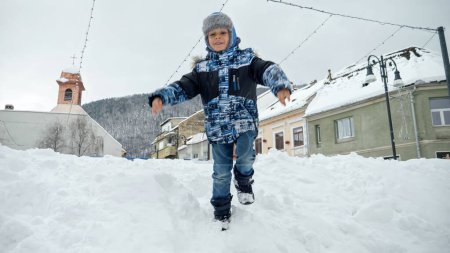 Foto de Feliz niño sonriente jugando y saltando en gran deriva de nieve en la calle. Bebé alegre, niños jugando en invierno, vacaciones de Navidad - Imagen libre de derechos