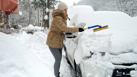 Foto de Mujer joven limpiando su coche con cepillo de nieve después de ventisca en el aparcamiento. Concepto de rutina diaria, cuidado del vehículo - Imagen libre de derechos