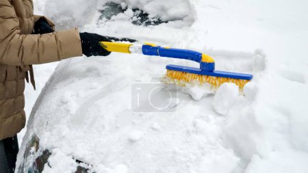 Foto de Primer plano de una joven limpiando la nieve de su coche con un cepillo. Concepto de invierno, mantenimiento de vehículos y tareas diarias - Imagen libre de derechos