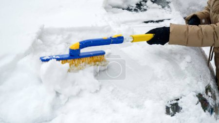 Téléchargez les photos : Gros plan d'une jeune femme balayant la neige de sa voiture par une journée enneigée. Concept d'hiver, d'entretien automobile et de tâches quotidiennes - en image libre de droit