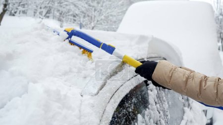 Téléchargez les photos : Gros plan d'une femme utilisant une brosse à neige pour enlever la neige du pare-brise de sa voiture après une forte chute de neige. Concept de temps hivernal, entretien de la voiture et préparation aux conditions enneigées - en image libre de droit
