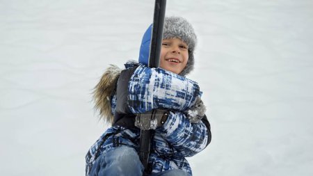 Téléchargez les photos : Joyeux garçon riant chevauchant sur la tyrolienne à l'aire de jeux publique couverte de neige après blizzard. Plaisir et joie pendant les vacances d'hiver, activité dans la neige - en image libre de droit
