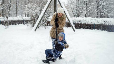 Téléchargez les photos : Mère heureuse avec son fils jouant sur une aire de jeux lors d'une journée d'hiver enneigée et chevauchant sur une tyrolienne. Plaisir et joie pendant les vacances d'hiver, jeu en famille, activité dans la neige - en image libre de droit