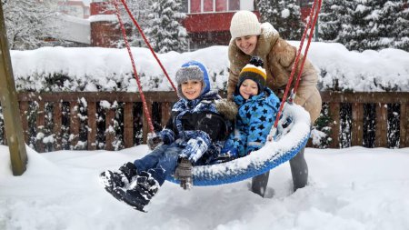 Foto de Dos hermanos con madre jugando felizmente en un columpio nevado en el parque público. Diversión invernal, vinculación familiar y actividad al aire libre para niños - Imagen libre de derechos