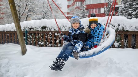 Téléchargez les photos : Amusement hivernal et rires pendant que deux frères se balancent sur une aire de jeux enneigée. Activité de plein air et plaisir en famille - en image libre de droit