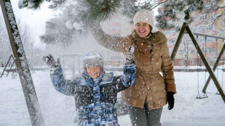 Photo for Laughing mother with son shaking tree branch and enjoying falling snow on heads. Fun and joy on winter holidays, kids playing outdoors, activity in snowfall - Royalty Free Image