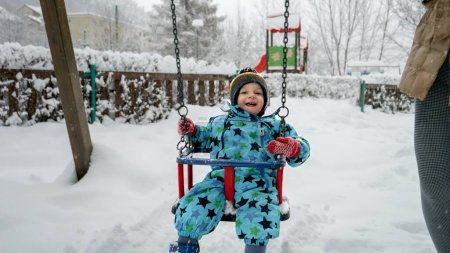 Téléchargez les photos : Un jeune garçon sourit en se balançant dans le parc enneigé par une froide journée d'hiver. Parfait pour transmettre la joie et la magie du jeu d'hiver pour les enfants - en image libre de droit