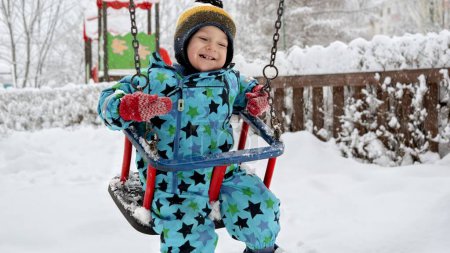 Téléchargez les photos : Bébé mignon garçon profitant d'une balançoire dans un parc d'hiver enneigé. Concept de plaisir et de joie en plein air, les enfants jouent dans les chutes de neige - en image libre de droit