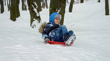 Téléchargez les photos : Joyeux garçon souriant descendant la colline enneigée sur des traîneaux en plastique. Concept de vacances d'hiver, les enfants s'amusent et jouent dehors dans la neige, vacances de Noël - en image libre de droit