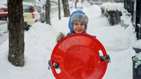 Foto de Niño alegre caminando con trineos de plástico en las nevadas en el parque de invierno. Concepto de niños divirtiéndose durante el invierno, vacaciones de Navidad y jugando al aire libre en la nieve - Imagen libre de derechos