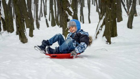 Photo for Young boy joyfully sledging down a snowy hill on his plastic sled. The snowy landscape, winter holidays and outdoor activities - Royalty Free Image