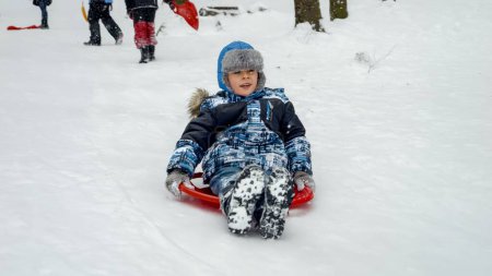 Photo for Cheerful young boy sledging down a snowy hill on his plastic sleds, exuding pure joy and enthusiasm. Holiday season and the fun children have in the snow - Royalty Free Image
