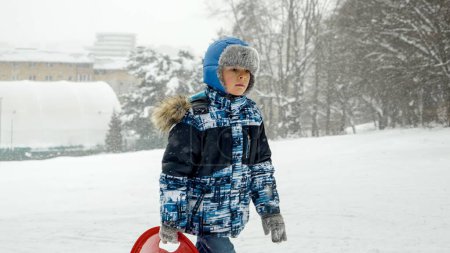 Téléchargez les photos : Un garçon souriant porte ses traîneaux sur une colline enneigée, prêt pour une descente passionnante. Joie et merveille du temps de jeu d'hiver - en image libre de droit