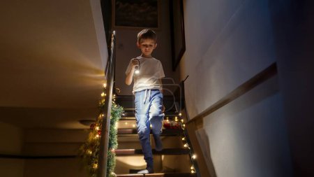 Foto de Emocionado niño esperando a Santa Claus en la víspera de Navidad bajando las escaleras de la casa con linterna. Niños celebrando vacaciones de invierno - Imagen libre de derechos