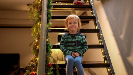 Téléchargez les photos : Petit garçon souriant mignon assis sur un escalier en bois décoré à la maison et attendant Noël ou le Nouvel An. Enfants célébrant les vacances d'hiver - en image libre de droit