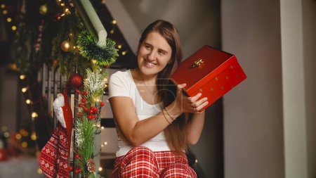 Téléchargez les photos : Joyeux rire et excité femme en pyjama tenant et secouant cadeau de Noël en boîte rouge du Père Noël tout en étant assis sur des escaliers en bois. Célébrations familiales pendant les vacances d'hiver - en image libre de droit