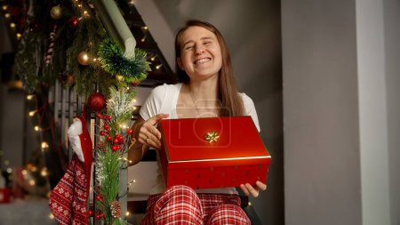 Photo for Happy cheerful woman in pajamas sitting on wooden stairs and holding Christmas present in gift box she received on winter holidays. - Royalty Free Image