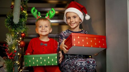Téléchargez les photos : Deux garçons joyeux en pyjama riant et excité après avoir reçu des cadeaux de Noël du Père Noël le Nouvel An assis sur des escaliers en bois à la maison. Célébrations familiales pendant les vacances d'hiver - en image libre de droit