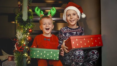 Foto de Retrato de dos chicos sonrientes en pijama sentados en las escaleras con regalos de Navidad y riendo en cámara. - Imagen libre de derechos