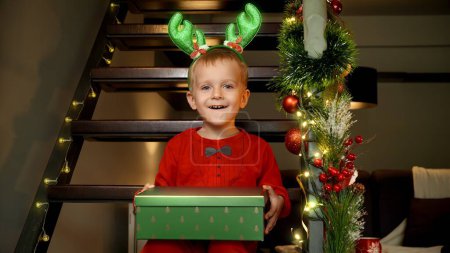 Foto de Retrato de niño lindo con astas divertidas sosteniendo regalo de Navidad y sentado en escaleras de madera en la casa en la celebración de Año Nuevo. - Imagen libre de derechos
