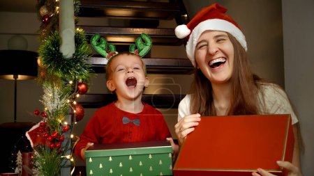 Téléchargez les photos : Joyeux garçon tout-petit qui rit avec sa mère tenant des cadeaux de Noël et des boîtes cadeaux du Père Noël et souriant à la caméra. Célébrations familiales pendant les vacances d'hiver - en image libre de droit